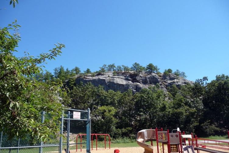 A photo of a prominent rock formation in the background and, in the foreground, a playground next to that rock. The rock is Rock Rimmon in Manchester, NH.